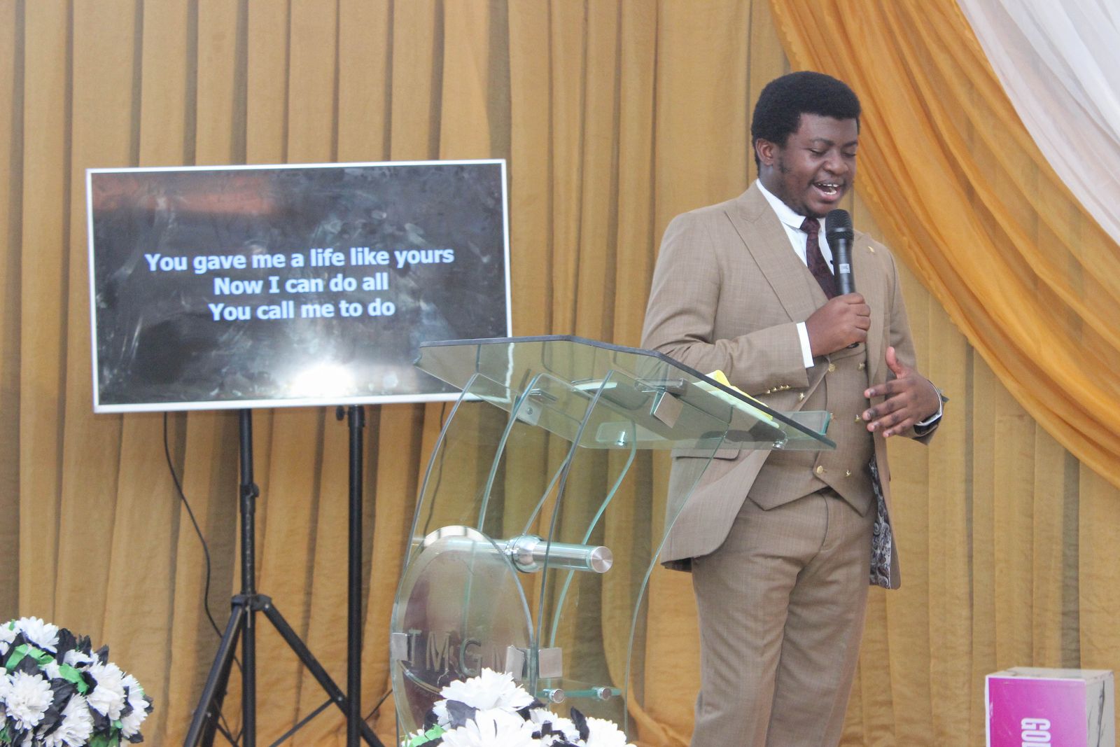 Our pastor in a brown suit teaching, with the scripture on the screen beside him